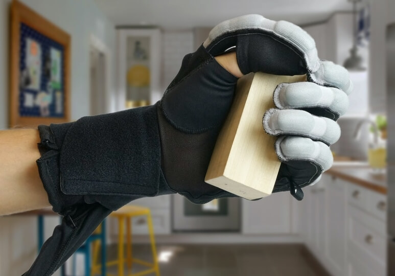 A hand wearing a soft robotic glove grips a small wooden block. 