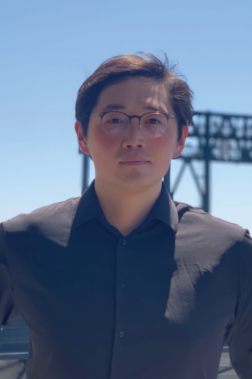A man in a black button down shirt and glasses stands in front of a blue, cloudless sky. 