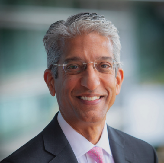 A man with white/gray hair and frameless glasses smiles in front of a blurry background.
