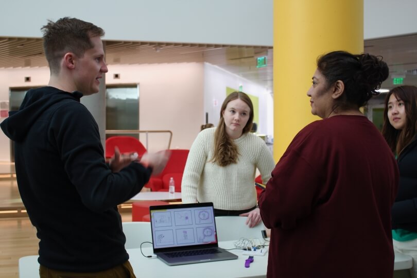 A man and a woman chat in front of a table with two female students behind it and a laptop and purple watch on the table
