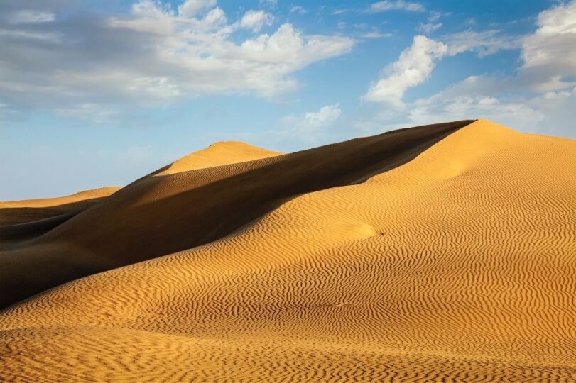 Golden sand dunes with wavy patterns under a blue sky with few clouds.