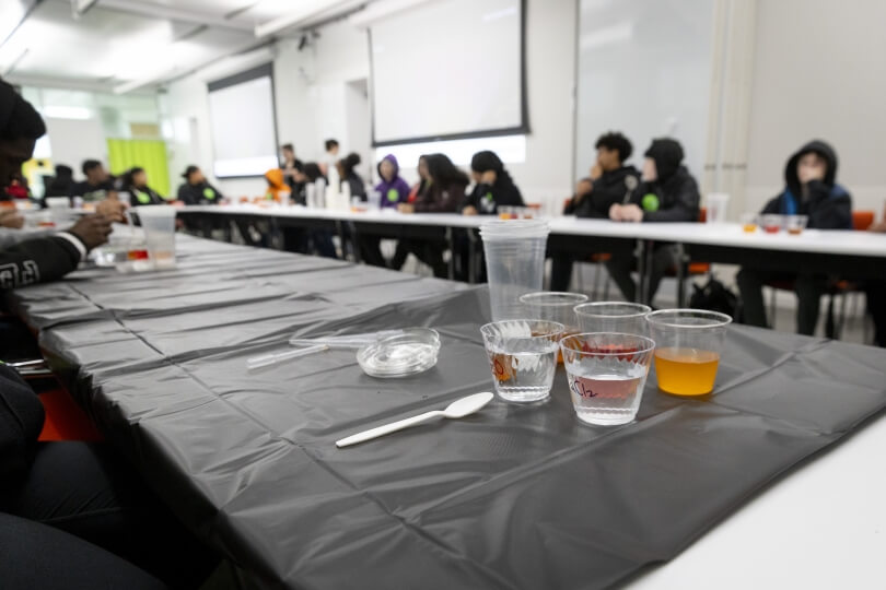 A group of students sit along tables behind a collection of plastic cups containing liquid