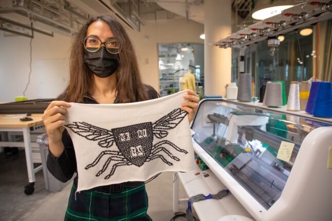Vanessa Sanchez holds up a piece of white knit fabric with the Harvard Microrobotics Lab logo. It is an insect with the Harvard shield as the body.