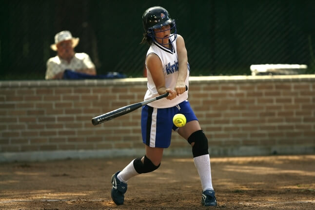 Woman in a white sleeveless jersey hits a yellow softball with a black baseball bat.