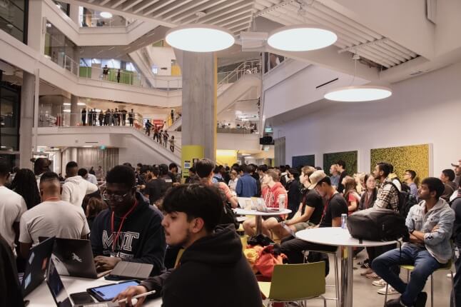A large crowd of students sitting at tables and standing on stairwells at HackHarvard 2023