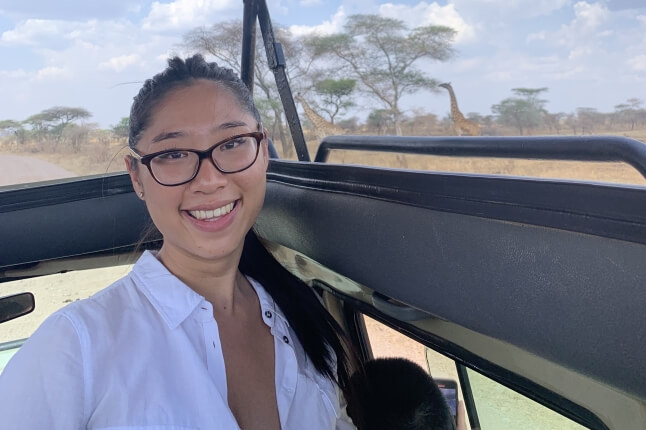Michelle Doan, A.B. ‘25 on a safari with a giraffe eating from a tree behind her