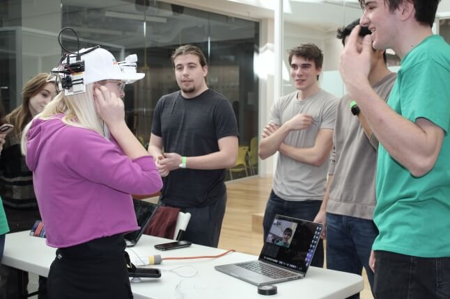 Four students stand behind a table with a woman wearing a white hat testing their device