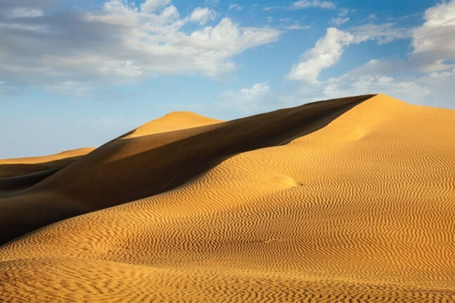 Golden sand dunes with wavy patterns under a blue sky with few clouds.